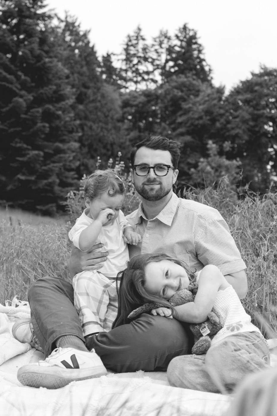 Jake Meaney sitting on a blanket in a summer meadow with his two young daughters resting against him.