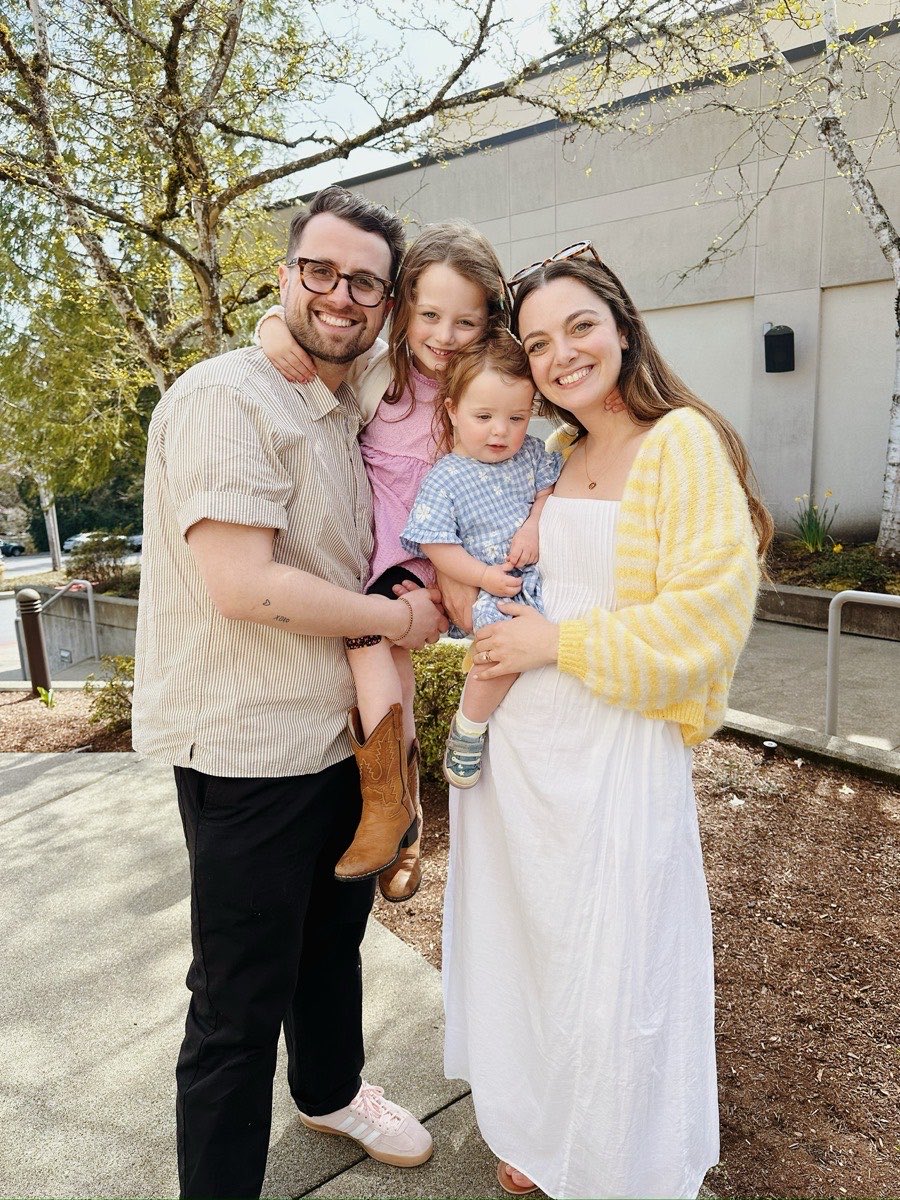 Jake Meaney outdoors on a spring day with his wife and their two young daughters, all of them smiling at the camera.