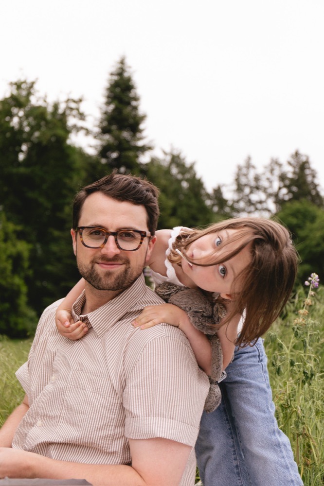 Jake Meaney in a field, in glasses and a striped shirt, with one of his daughters leaning over his shoulder.